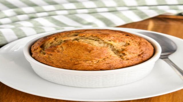 Closeup of banana bread baked in a small white ramekin set on a white plate that in turn is set on a wooden cutting board with a green and white striped napkin in the background.