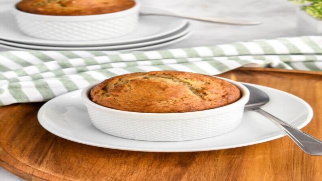 A single serve banana bread in a ramekin set on a white dish with a spoon next to it which in turn is set on a wooden cutting board with a second ramekin of banana bread showing in the background and a green and white striped napkin.